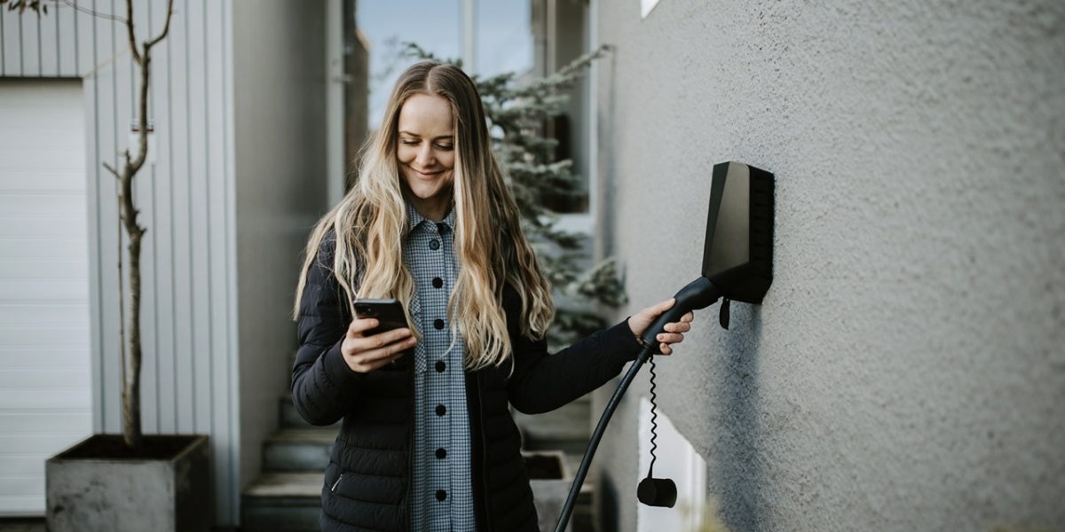 Ung kvinne med langt lyst hår styrer sort elbillader på grå murvegg med en smarttelefon