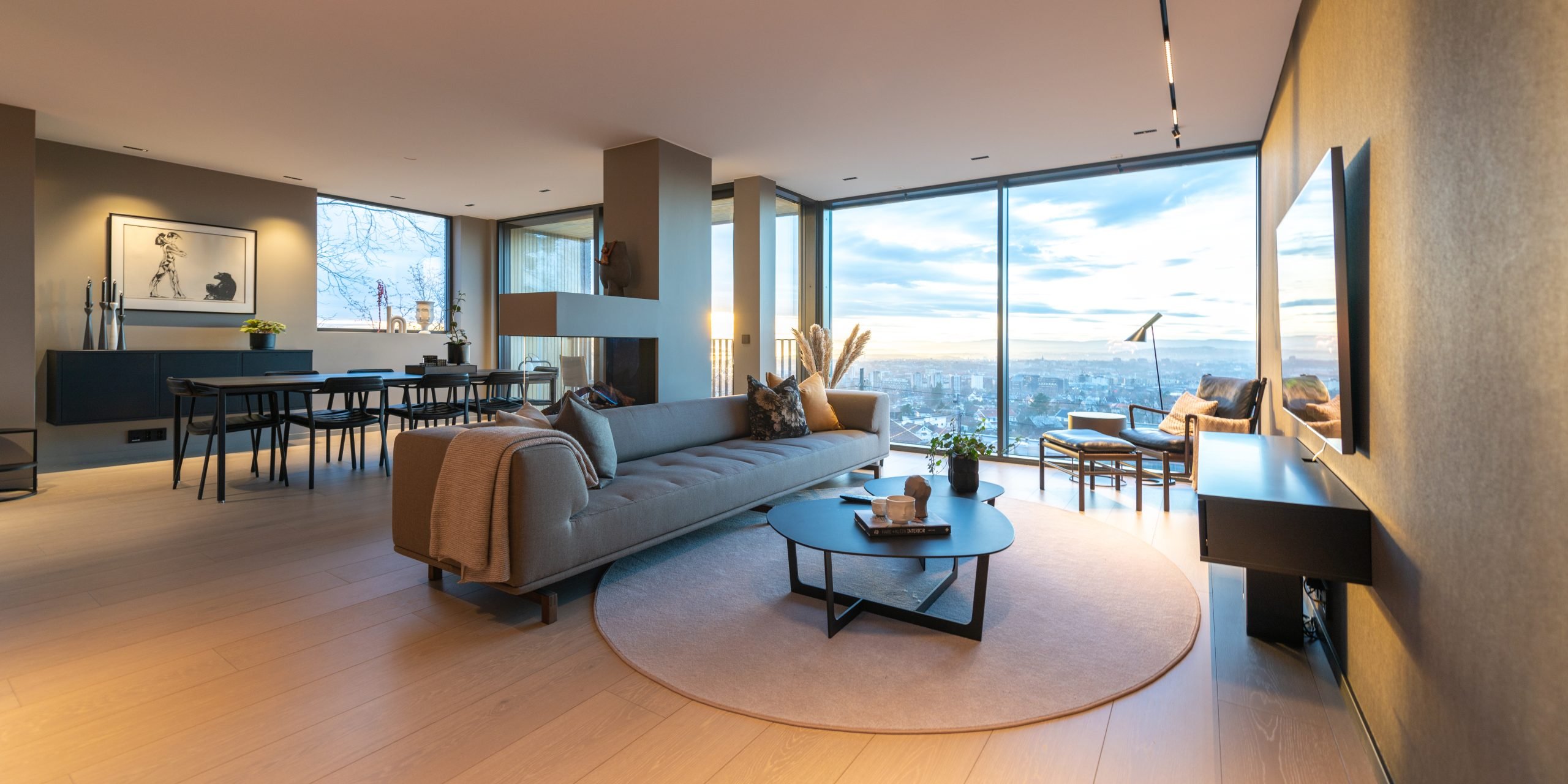 Stor moderne stue med grå sofa, stor spisegruppe og store vindusflater med utsikt. 