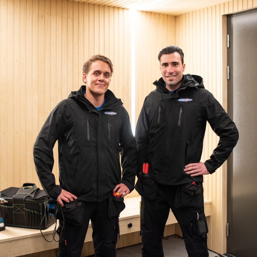 To elektrikere med Norgeseliten jakke foran et opplyst inngangsparti