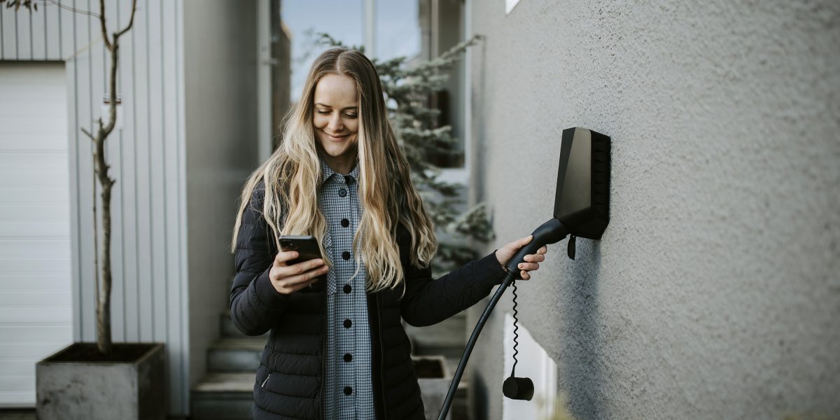 Dame som står foran inngangspartiet til et hvitt hus med glassdør og setter i elbilladeren i veggen mens hun smiler og ser ned i mobiltelefonen