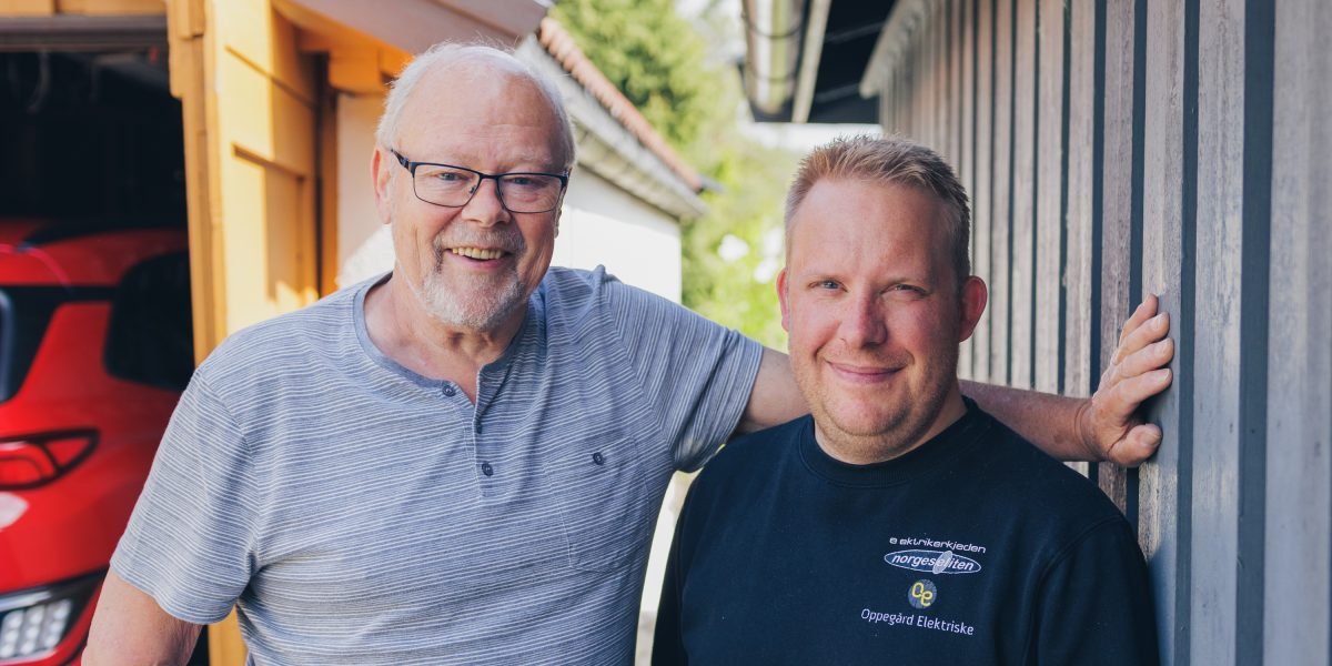 Elbileier Dag Røneid og elektriker Anders Bakke står smilende side om side foran garasje med rød elbil parkert