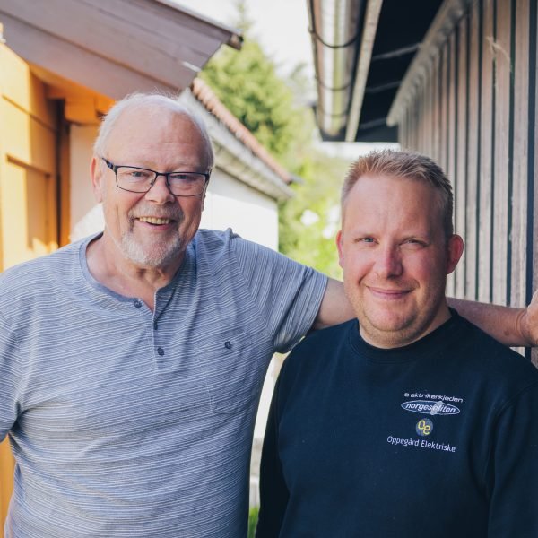 Elbileier Dag Røneid og elektriker Anders Bakke står smilende side om side foran garasje med rød elbil parkert