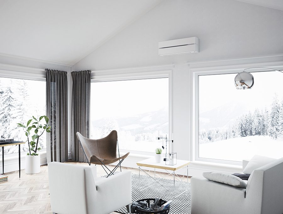 hvit varmepumpe som er montert på en hvit vegg over to store vinduer, og en grå sofa og to stoler i forgrunnen med en grønn plante og snølandskap utenfor boligen.