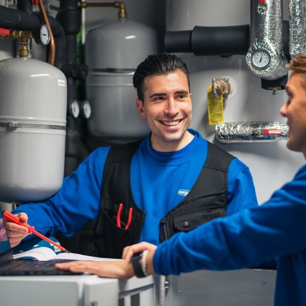 To Norgeseliten-elektrikere i blå gensere som smiler til hverandre og noterer ned på et ark i et rom med elektrisk anlegg.