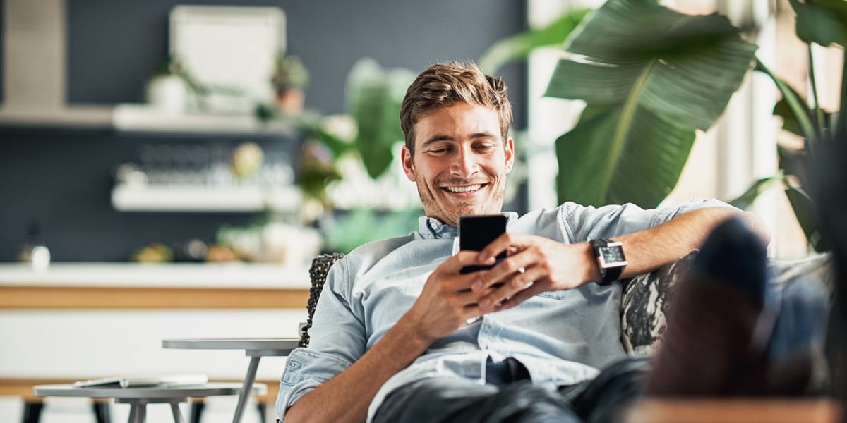 mann som smiler og bruker mobilen mens hans sitter i en sofa med en grønn plante foran seg