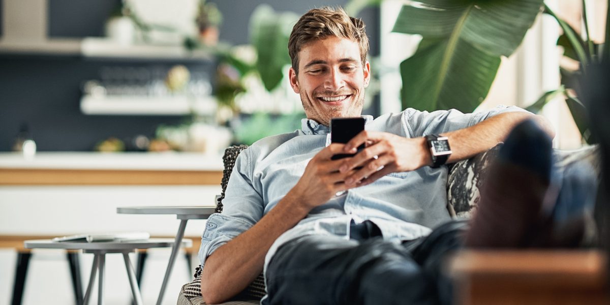 mann som smiler og bruker mobilen mens hans sitter i en sofa med en grønn plante foran seg