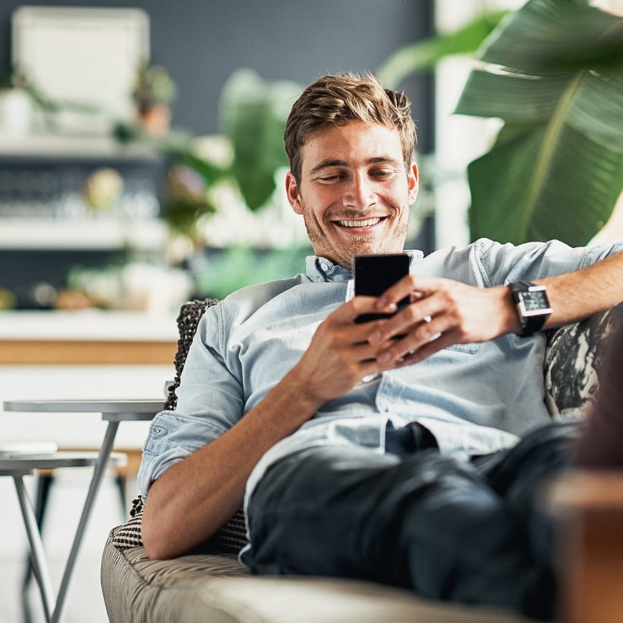 mann som smiler og bruker mobilen mens hans sitter i en sofa med en grønn plante foran seg