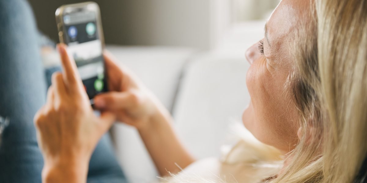 En dame med blondt hår som ligger på sofaen og smiler og holder en mobiltelen med en app-løsning fra porttelefon-leverandøren Urmet