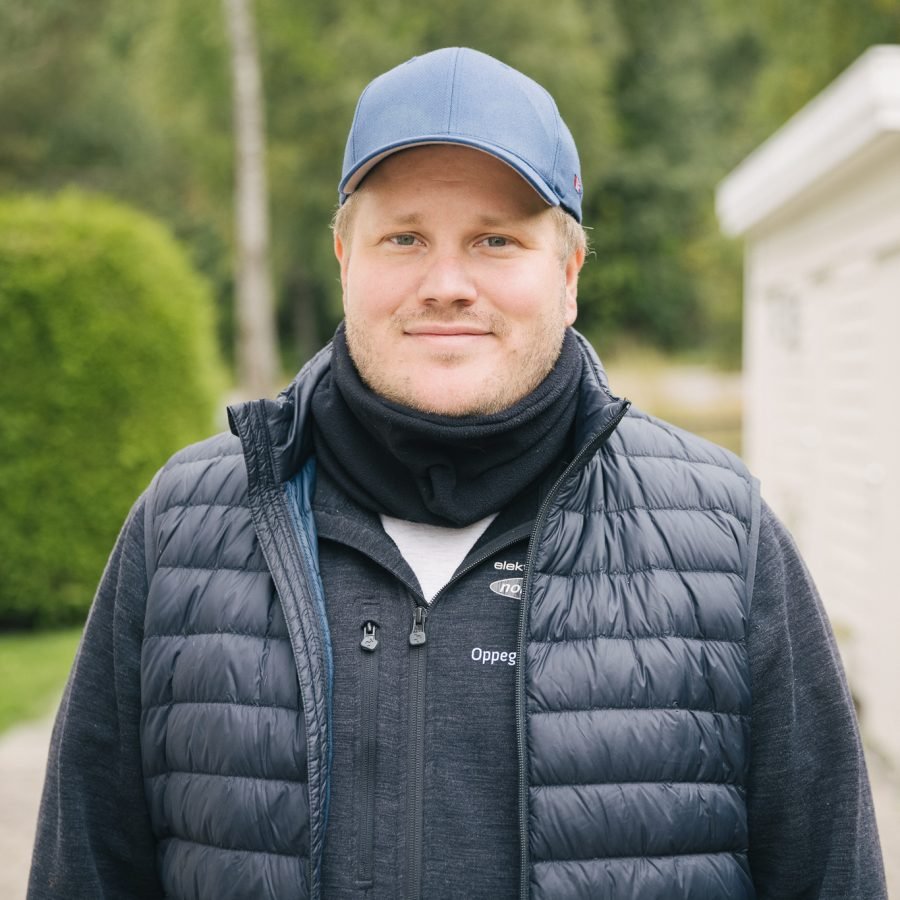 En Norgeseliten-elektriker fra Oppegård Elektriske med sort genser og boblevest, og blå caps som står ved inngangen til en bolig og smiler inn i kameraet