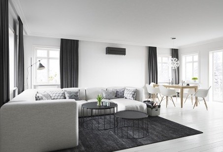 Stor lys stue med hvite vegger, hvit hjørnesofa og mørkegrå gardiner og gulvteppe med sort varmepumpe som gir enovastøtte på veggen.