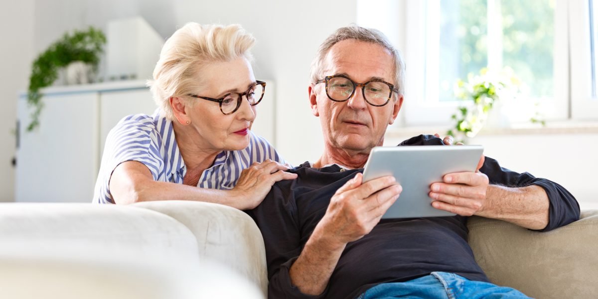 Eldre mann sitter i sofa og viser noe på et nettbrett til sin kone.