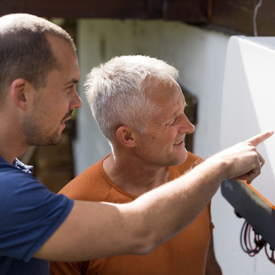 Elektriker i blå t-skjorte og mann i oransje skjorte inspiserer en vekselsretter montert på husvegg.