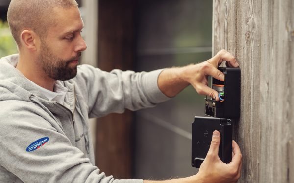 ung elektriker med grå genser monterer veggfeste til elbillader på brun husvegg